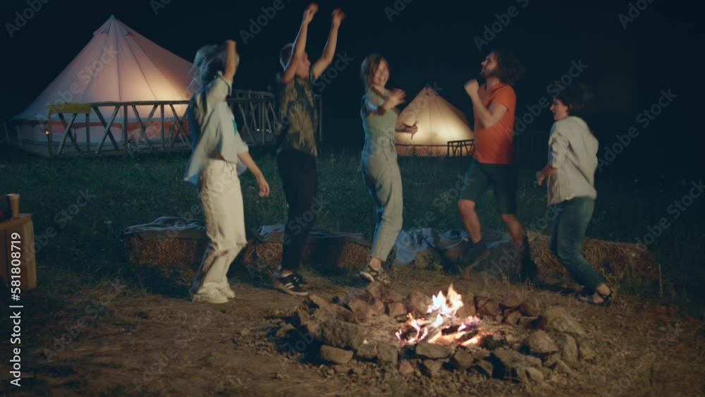 In the middle of campsite beside the fire stake group of students have party time in the night they dancing and moving excited all together In the middle of campsite beside the fire stake group of students have party time in the night they dancing and moving excited all together
