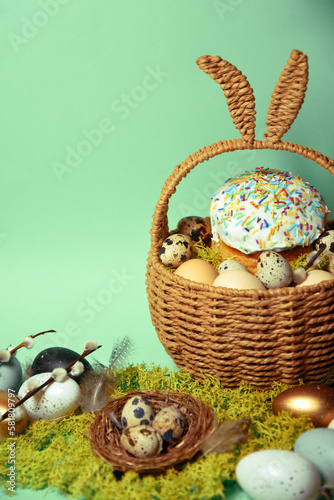 Easter decorations in a basket with rabbit ears. Easter cake and eggs on moss  quail eggs and feathers on mint green background. pussy willow Place for text.