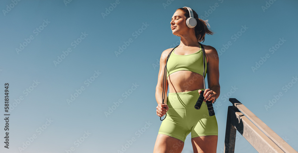 Female athlete in her 30s outdoors taking a break from her cardio workout, holding a skipping rope and wearing sportswear - obrazy, fototapety, plakaty 
