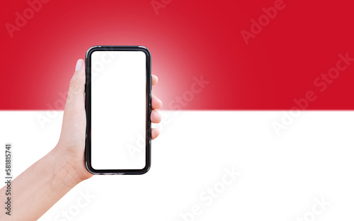 Male hand holding smartphone with blank on screen, on background of blurred flag of Monaco. Close-up view.