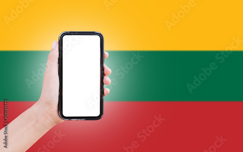 Male hand holding smartphone with blank on screen, on background of blurred flag of Lithuania. Close-up view.