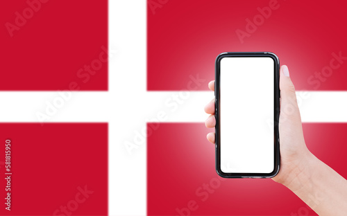 Male hand holding smartphone with blank on screen, on background of blurred flag of Denmark. Close-up view.
