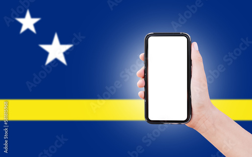 Male hand holding smartphone with blank on screen, on background of blurred flag of Curacao. Close-up view.