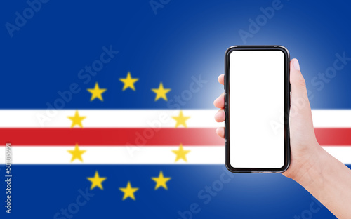 Male hand holding smartphone with blank on screen, on background of blurred flag of Cape Verde. Close-up view.