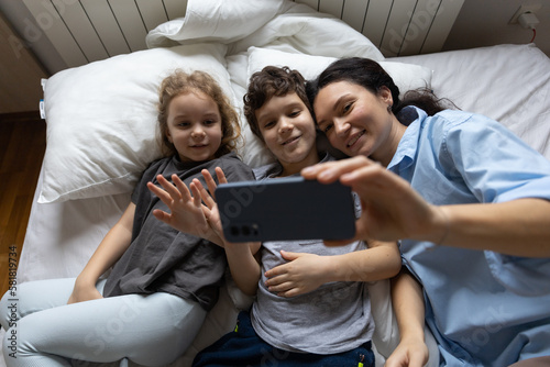 Happy caucasian mother and 2 kids have online video call with father or grandmother mobile phone when lying on bed at home. the concept of modern communication.lifestile. top view