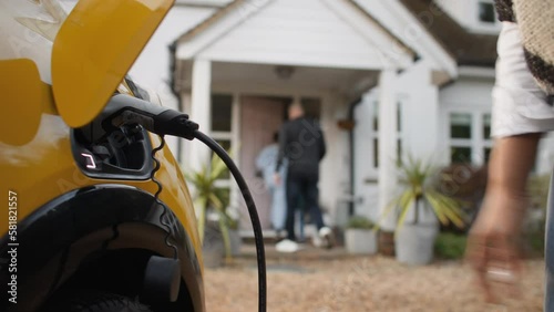 Close up of woman plugging electric charger into car with multi ethnic family walking into home
