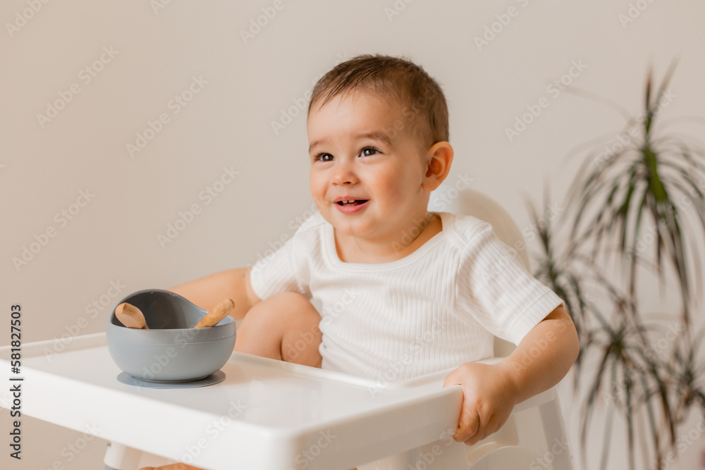 portrait of a cute baby in a light bodysuit sitting in a high chair eating from children's dishes. The concept of baby food portrait of a cute baby in a light bodysuit sitting in a high chair eating from children's dishes. The concept of baby food