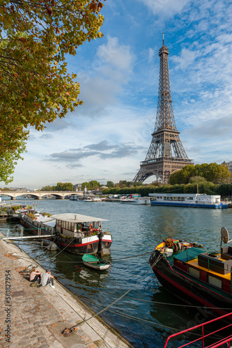 View of the Eiffel Tower with the River Seine at a sunny day in Autumn. House and Restaurantsboats are towed at the River bank. A Couple is enjoying the view.