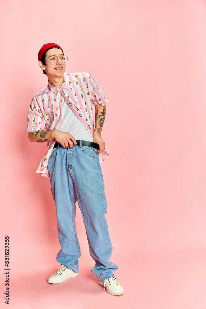 Fototapeta premium Full length photo of stylish guy with tattoo fastening his belt and looking away over pink background. Concept of fashion, youth, student life
