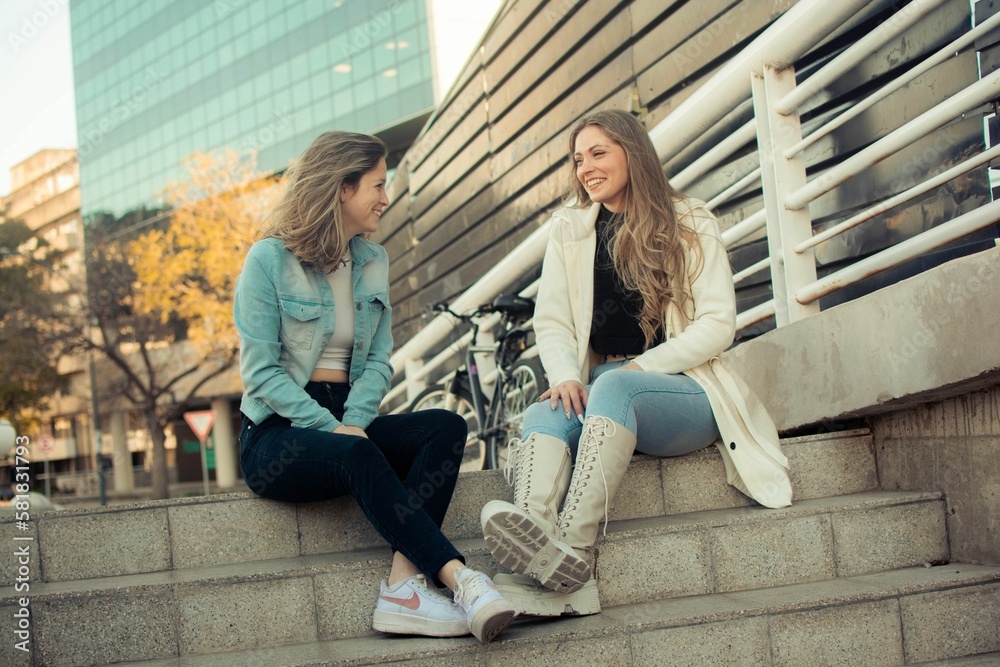 Pair of young stylish female friends sitting together outdoors in the city