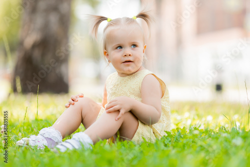 a happy little blonde girl in a yellow dress is sitting on the green grass in the park in summer. Earth Day. Children Protection Day