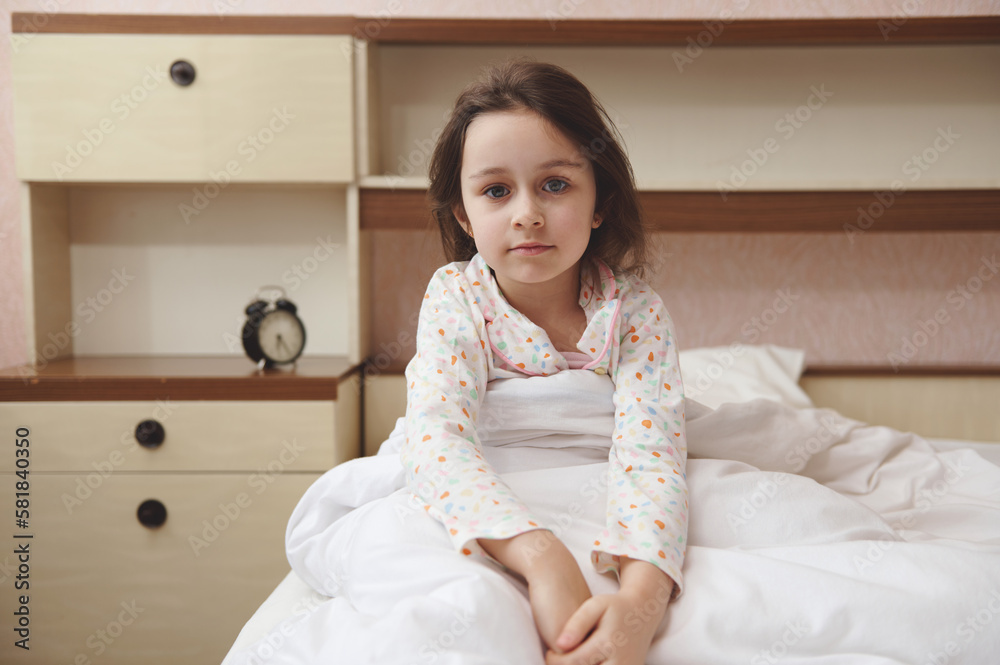 Portrait of a Caucasian beautiful child girl in pajamas, sitting in the bed at her cozy bedroom. Children. Lifestyle. Bedtime. Morning routines. The concept of going to bed, waking up and relaxing Portrait of a Caucasian beautiful child girl in pajamas, sitting in the bed at her cozy bedroom. Children. Lifestyle. Bedtime. Morning routines. The concept of going to bed, waking up and relaxing