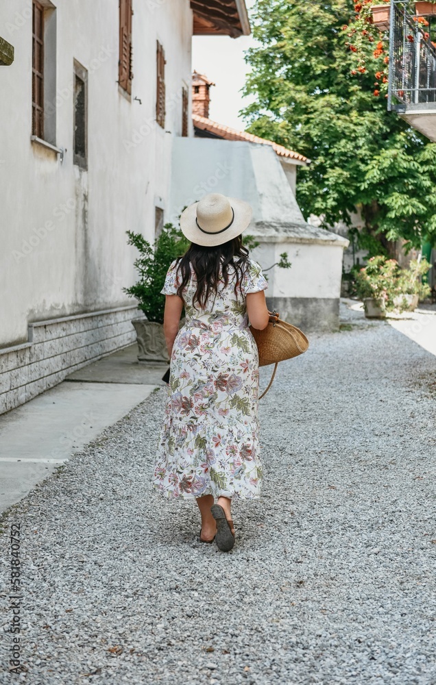 Vertical shot of a young female tourist in a floral dress and sunhat walking in an old town