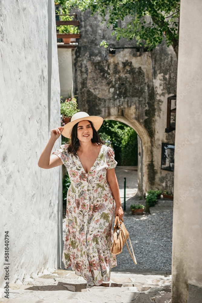 Vertical shot of a young female tourist in a floral dress and sunhat posing in an old town