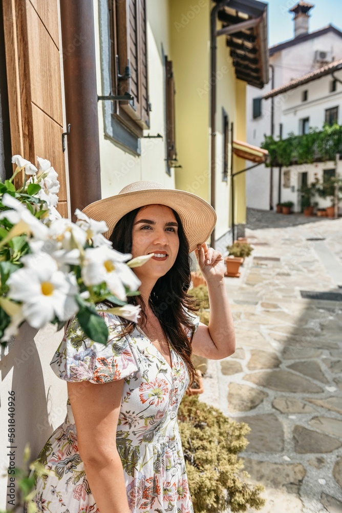 Smiling young woman in a floral sundress and a straw hat leaned against the wall in Smartno