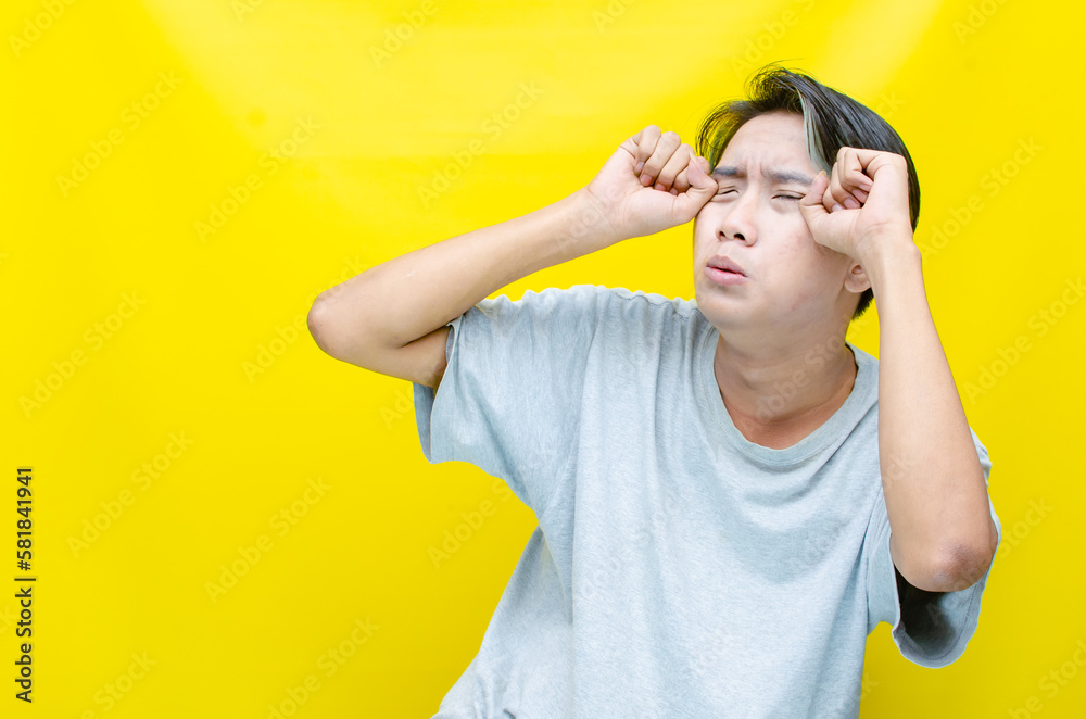 Sad Young Asian man wearing grey t-shirt with crying face expression isolated over yellow background. Crying face expression of Young Indonesian man with hand gesture. Sad Young Asian man wearing grey t-shirt with crying face expression isolated over yellow background. Crying face expression of Young Indonesian man with hand gesture.