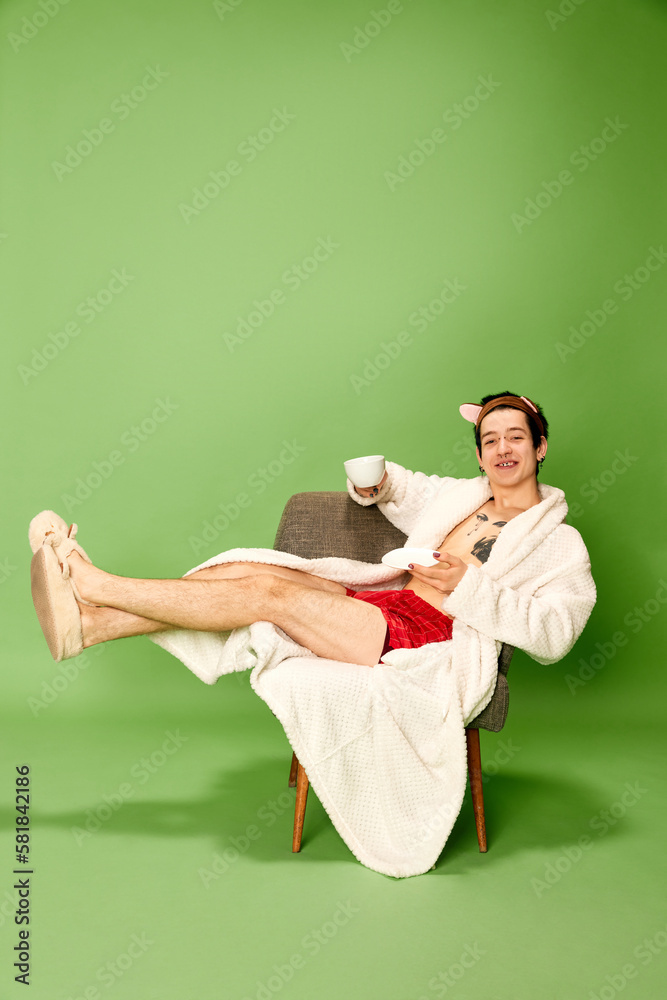 Young man wearing female bathrobe and slippers sitting on chair with cup of tea over green background. Concept of vacation, self love and self care, weekend, rest, relax