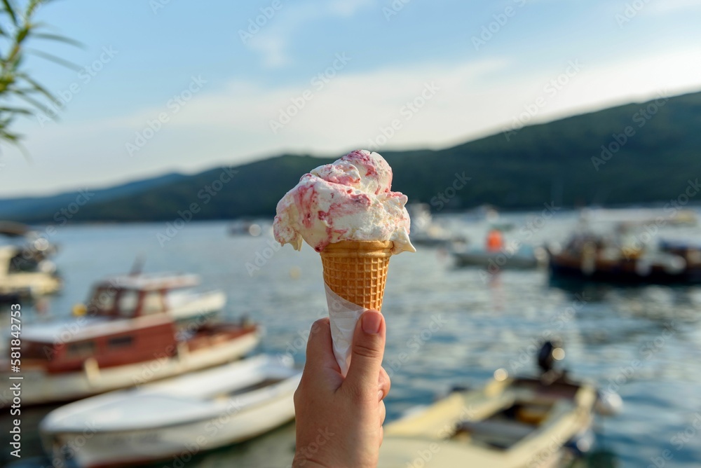 Naklejka premium Selective focus shot of a hand holding an ice cream in a cone with moored boats in the sea
