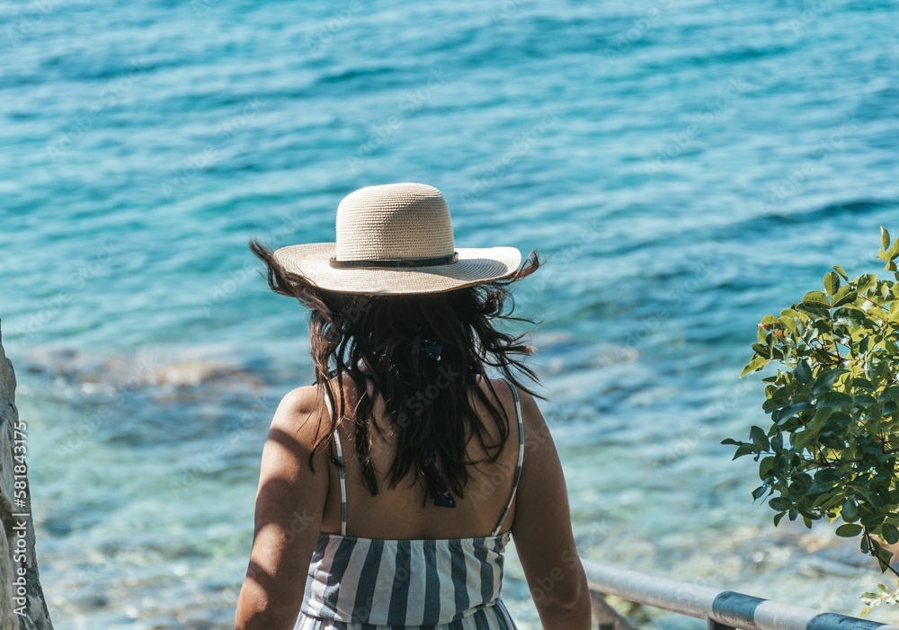 Woman wearing a striped sundress and hat, walking toward the beach with turquoise sea