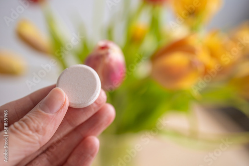 round large tablet, vitamin in the girl's hand, against the background of yellow tulips, indoors. Flu and cold season, new virus strain. Lack of vitamins in spring and winter, uncontrolled medication