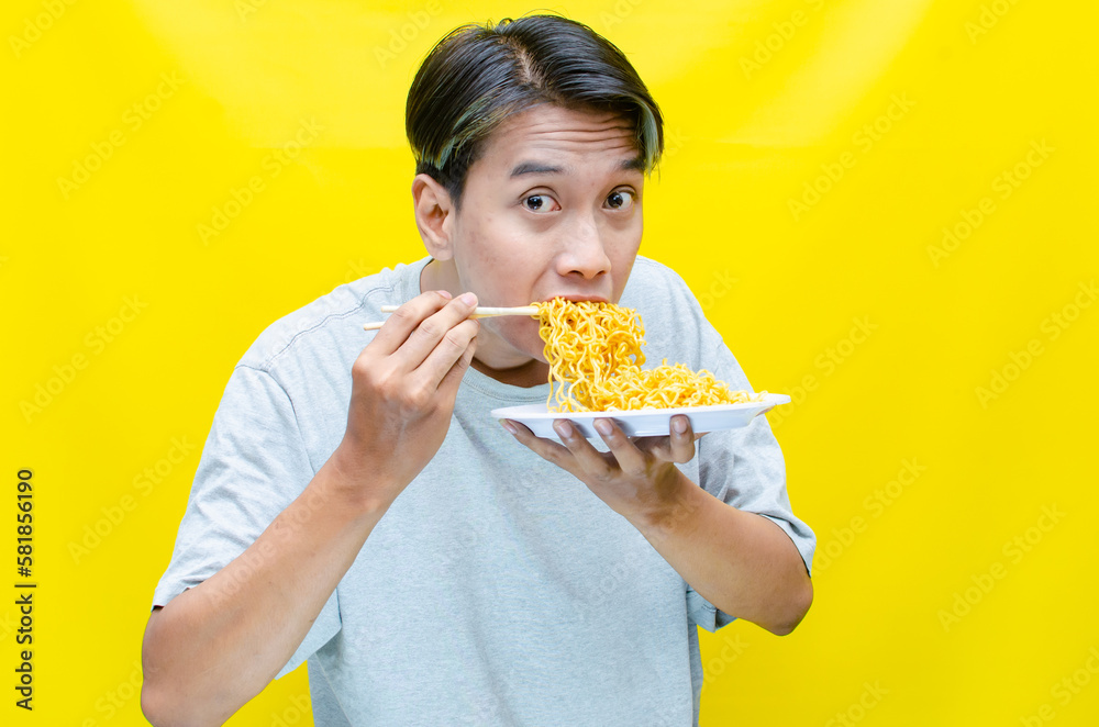 Happy euphoric asian young man in grey tshirt eating instant fried noodle using bamboo chopstick isolated over yellow background. stylish asian man with peek a boo hair eats instant noodle Happy euphoric asian young man in grey tshirt eating instant fried noodle using bamboo chopstick isolated over yellow background. stylish asian man with peek a boo hair eats instant noodle