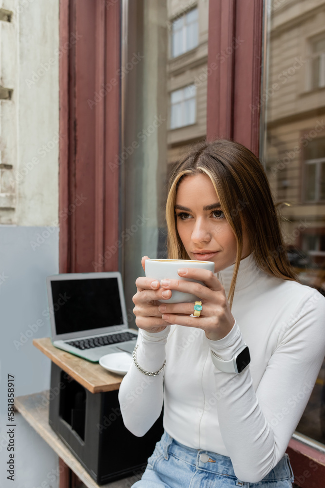 young woman drinking coffee while sitting in outdoor cafe next to laptop with blank screen in Vienna. young woman drinking coffee while sitting in outdoor cafe next to laptop with blank screen in Vienna.