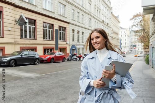 Photography pleased woman in stylish trench coat holding smartphone and laptop while walking on street in Vienna