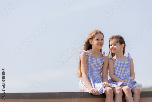 two happy little girls are sitting on a wooden pier on a city lake in summer. Blue sky. Earth Day. Children's Day.Space for text