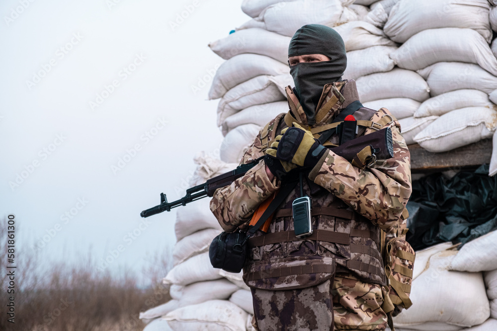 Obraz premium Soldier with weapon in military uniform stands next to the barricades made from sandbags and anti-tank hedgehog barriers. Military man on the roadblock. Combatant in full ammunition. Concept of war