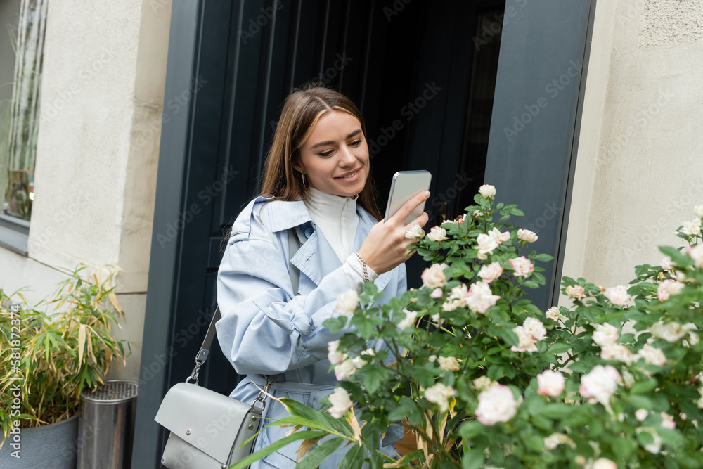 happy young woman in blue trench coat taking photo of green bush with blooming flowers on street in Vienna.