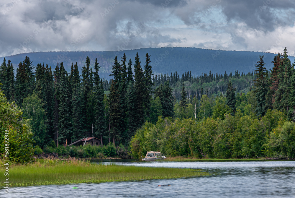 Fairbanks, Alaska, USA - July 27, 2011: Chena River wide landscape with green forest, mountain in back under blueish cloudscape. Small boat and wooden hut at far end shoreline
