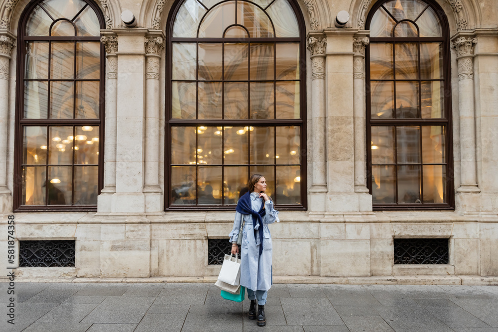 young and stylish woman with scarf on top of blue trench coat holding shopping bags near historical building in Vienna.