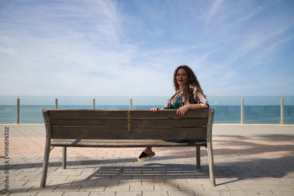 beautiful young woman, blonde with curly hair and blue eyes is sitting on a bench looking out to sea. The woman has the Atlantic Ocean in the background.