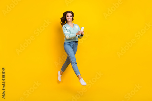 Full length photo of sweet cute woman wear turquoise cardigan jumping high texting device isolated yellow color background