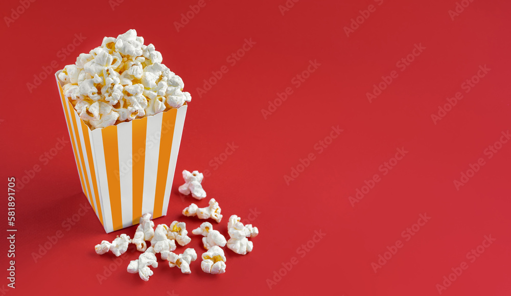 Yellow white striped carton bucket with tasty cheese popcorn, isolated on red background. Box with scattering of popcorn grains. Fast food, movies, cinema and entertainment concept.
