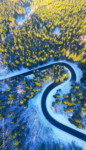 Asphalt road serpentine in snowy wintertime. Cold winter and sunny day above forest road with illuminated trees by rising sun. Aerial view from drone.