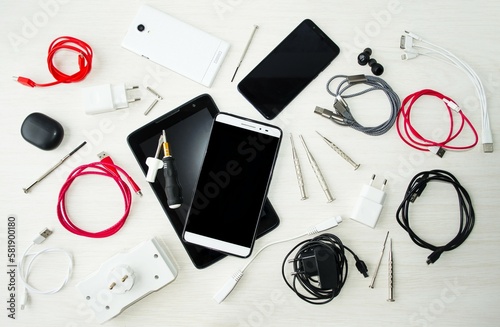 On the table on a light background in a chaotic manner are a tablet, a phone, headphones, a charger and adapters for electronic equipment.  The concept of accessories for equipment, repair products