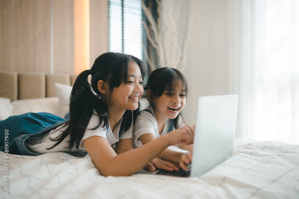 Fototapeta premium Sibling Asian girl child on the bed and learning online on a laptop Internet. Virtual class lesson on video during self isolation at home. Distant remote video education. Modern school study for kids