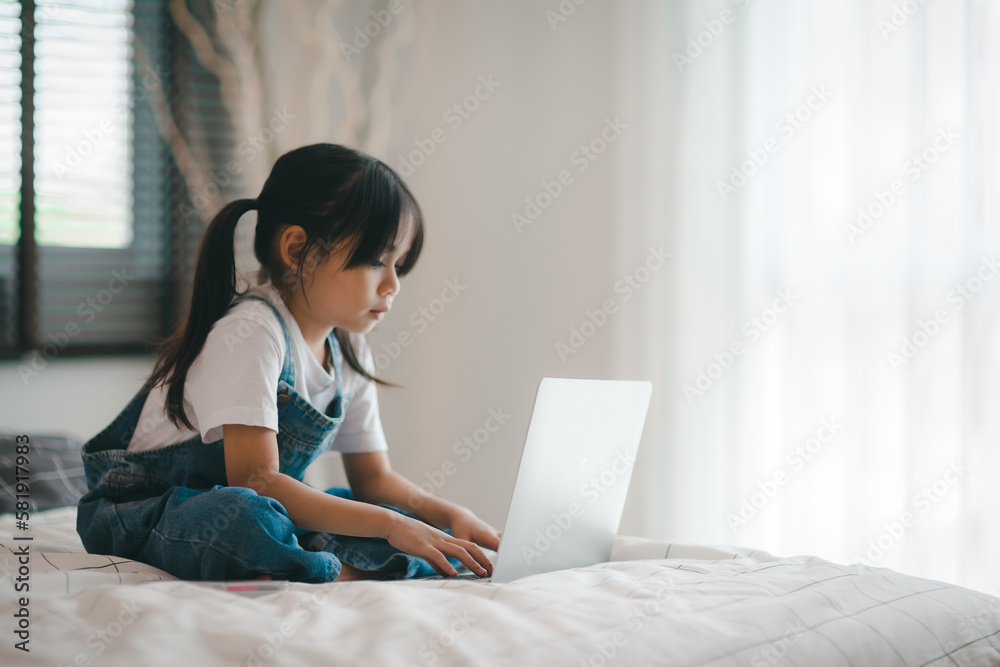 Asian girl child sitting in bed and learning online on laptop Internet. Virtual class lesson on video during self isolation at home. Distant remote video education. Modern school study for kids Asian girl child sitting in bed and learning online on laptop Internet. Virtual class lesson on video during self isolation at home. Distant remote video education. Modern school study for kids