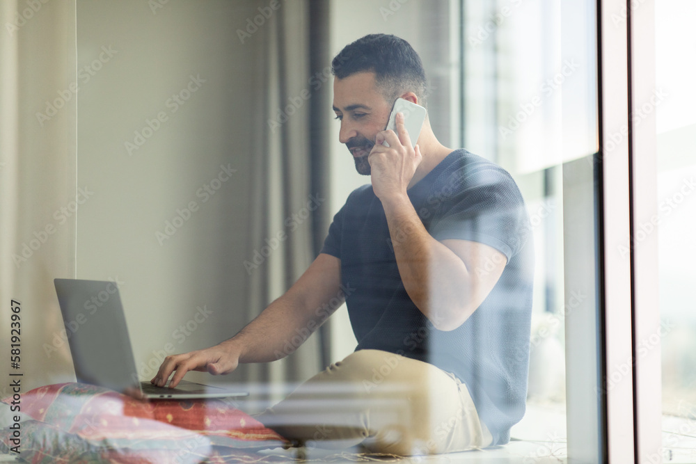 Fototapeta premium Middle aged man talking on cellphone and using laptop, sitting on floor near window at home, free space. Male working on pc