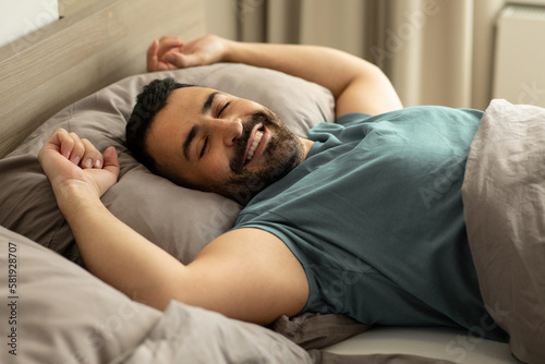 Happy relaxed man lying in comfy bed, enjoying good morning in bedroom, stretching his body after waking up