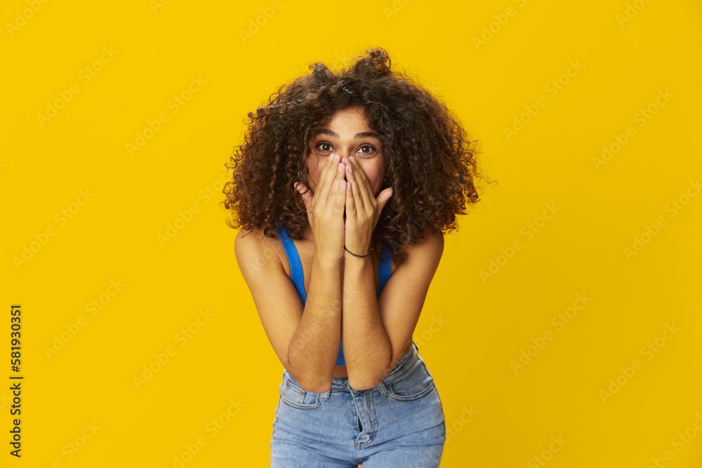 Woman with curly afro hair in a blue t-shirt on. yellow background signs with her hands, look into the camera, smile with teeth and happiness, copy space Woman with curly afro hair in a blue t-shirt on. yellow background signs with her hands, look into the camera, smile with teeth and happiness, copy space