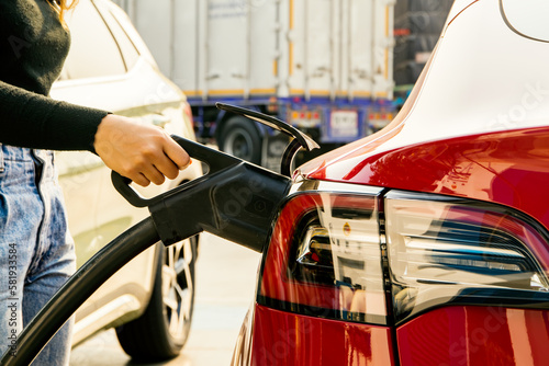 Young female EV or Electric car driver's hand holding DC power plug to her red electric car battery socket in gas station : Clean energy vehicle Alternative energy is becoming popular concept. photo