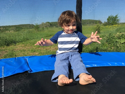 curly cute bold smiling little funny boy dancing with his hands up while sitting on a trampoline