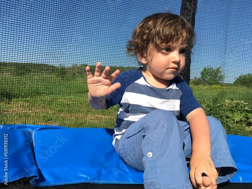 curly child boy welcomes raise his hand and waving his palm looks attentively sits on a trampoline