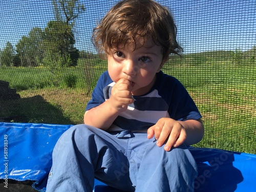 cute curly baby boy eating fingers wide open eyes sitting on a trampoline