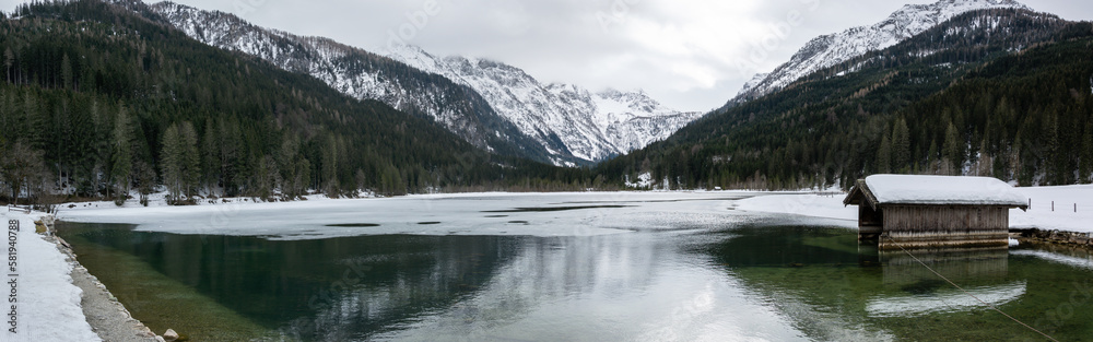 Fototapeta premium Panorama of the Jagersee lake in the Austrian mountains. Frozen water of a lake in the snowy alps with a wooden hut on the water.