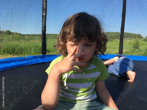 cute curly child boy rubbing one eye with his finger sits on a trampoline