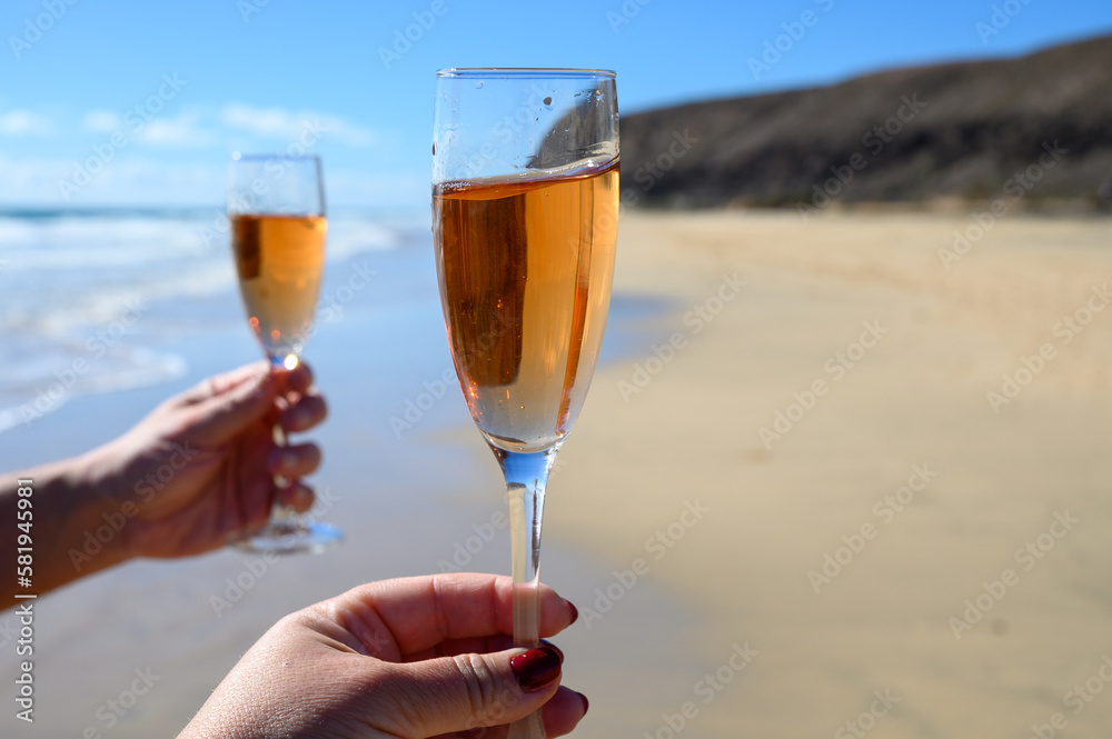 Beach party, hands with glass of rose champagne or cava sparkling wine, view on white sandy tropical beach and blue ocean water, romantic vacation, winter sun on Fuerteventura, Canary, Spain