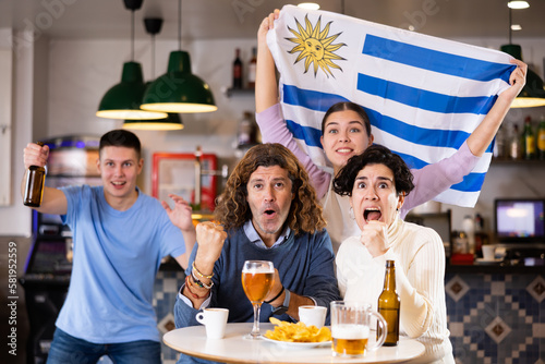 company of emotional young adult sports fans waving the flag of Uruguay and supporting their favorite team with beer in the pub. High quality photo